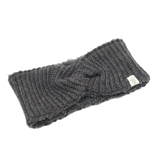 The Tuk Headband is a stylish dark grey accessory, featuring a unique twist design in the center and displayed flat against a white background. Hand-knit from soft merino wool, it includes a label tag on the side for added detail.