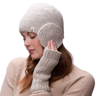 A person with long, reddish-brown hair is wearing a cream-colored knit beanie and matching Diagonal Knit Handwarmers, handmade in Nepal. They are holding the left handwarmer to their ear while looking down and smiling slightly. They are also dressed in a beige sweater. The background is white.