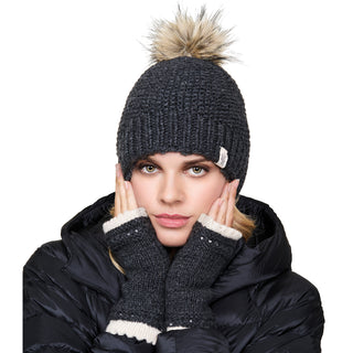 A person wearing a black knit beanie with a faux fur pom-pom, a black puffer jacket, and matching Les Handwarmers—black fingerless gloves with white trim made from soft merino wool—is holding their gloved hands up to their face against a white background.
