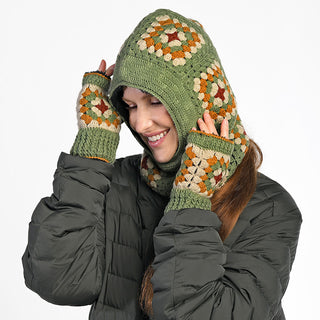 A person smiles with eyes closed against a plain white background, wearing green Ryan Handwarmers and a matching green hooded scarf with a multicolored granny square pattern, paired with a dark quilted jacket.