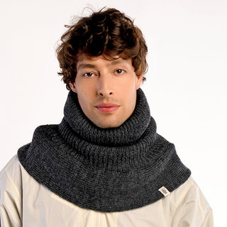 A person with short, wavy brown hair wears the thick, dark gray Hank Neckwarmer—handmade in Nepal—over a light beige top, facing forward against a plain white background.