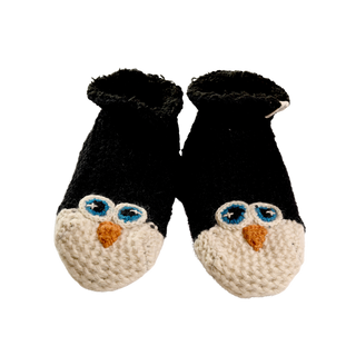 A pair of Penguin Slippers, featuring black and white, 100% wool knitted baby booties with blue and white eyes and an orange beak, designed to resemble penguins, set against a black background.