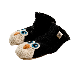 A pair of black children's slippers, hand-knit in Nepal from 100% wool, featuring adorable penguin faces stitched onto the toes. The penguin faces have large blue and white eyes and an orange beak. White tags are attached near the ankle opening for added charm.