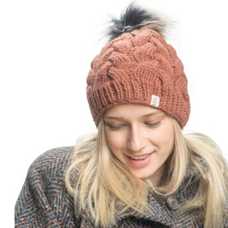 A person with long blonde hair is looking downward and smiling, wearing a patterned thick brown coat and the Boheme Cable Beanie w/ Faux Fur Pom, made from 100% Merino Wool. The background is white.