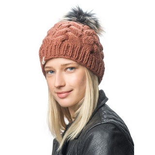 A person with long blonde hair is wearing a brown Boheme Cable Beanie with a black and white faux fur pom-pom on top. They are dressed in a black leather jacket and are posing in front of a white background, showcasing the cozy style of their 100% Merino Wool winter beanie.