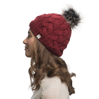 A woman with long, wavy brown hair is seen from the side, wearing a Boheme Cable Beanie w/ Faux Fur Pom in red. She is dressed in a light-colored, knitted sweater made of 100% Merino Wool with a subtle pattern. The background is plain white.