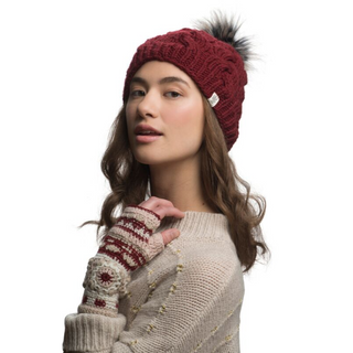 A woman with long brown hair is wearing a Boheme Cable Beanie w/ Faux Fur Pom in red and a beige sweater. She has fingerless gloves with a red and white pattern on her right hand and is posing against a white background.