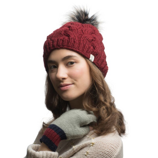 A person is wearing a Boheme Cable Beanie w/ Faux Fur Pom and a pair of light gray gloves with red and navy stripes at the cuffs. They have long, wavy brown hair and are dressed in a light beige sweater.