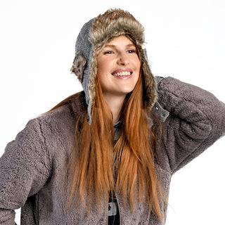 A person with long red hair wears a furry grey winter jacket and the Fern Hat—a handmade in Nepal grey trapper hat with faux fur trim—smiling with one arm behind their head against a plain white background.