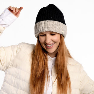 A woman with long red hair smiles and raises one arm against a white background while wearing the Cliff Hat, a black and beige wool beanie with faux shearling lining, along with a cream puffer jacket.