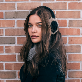 A woman with long brown hair stands against a brick wall. She is wearing Cable Knit Adjustable Earmuffs With Faux Fur, a black coat, and a gray knitted scarf. She looks slightly to the side with a neutral expression.