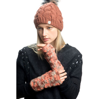 A person with long blonde hair is wearing a Boheme Cable Beanie with a faux fur pom-pom, a black sweater with lace-up sides, and matching fingerless gloves. The individual appears to be looking slightly downward while holding their chin with a gloved hand.