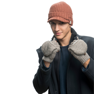 A person wearing a brown knit cap, Bryant Fingerless Gloves with Flap, a dark jacket, and a blue sweater stands against a plain white background. They have their mitten-covered fists near their face and are smiling slightly.