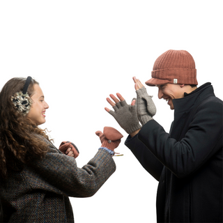 Two people, one wearing a brown beanie and gray Bryant Fingerless Gloves w/ Flap, the other in earmuffs and a coat, face each other smiling. They hold winter gloves and appear to be about to put them on or are playfully interacting with the mittens.