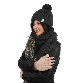 A person dressed in winter attire, wearing a black knitted hat with a pom-pom, a matching black scarf, a colorful woven scarf, a black leather jacket, and cozy Bryant Fingerless Gloves w/ Flap. The individual is smiling and crosses their arms in front of their body.
