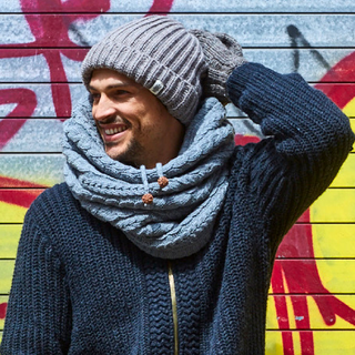 A man in a Lou Neckwarmer and hat is standing in front of a graffiti wall.