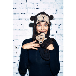 A woman wearing a Cheeta Monkey Hatscarf and matching mittens stands in front of a white brick wall, smiling at the camera.