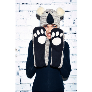 A person wearing a Koala Hatscarf and large, paw-like mittens is holding up their hands to frame their face, smiling at the camera against a white brick wall background.