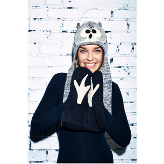 A smiling woman wearing a whimsical merino wool Chouette Hatscarf and holding hand-knit gloves against a white brick wall background.