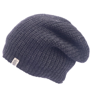 The Cardigan knit slouch, a black ribbed beanie, is shown on a white background. Handmade in Nepal from 100% wool, it offers a slouchy fit and includes a small rectangular side tag with a logo.