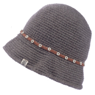 The Emily Sun Hat is a gray crochet hat that boasts a soft, textured appearance and features a charming brown braided band adorned with small white buttons encircling it. It also includes a small fabric logo tag on its side, adding to its handcrafted appeal.