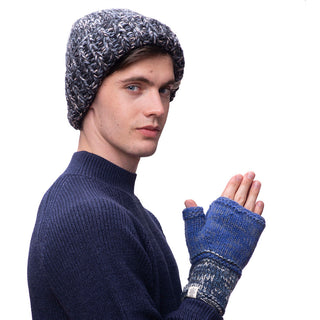 A person wearing a Marbled Rib Fold Beanie in a knitted blue-gray style and knitted fingerless gloves, both handmade in Nepal from soft Merino wool, stands against a white background. The person is dressed in a dark blue sweater and seems to be clapping or holding their hands close together in front of their face.