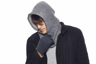 Wearing a dark coat and the Roll Hood, a grey hoodie with a wool hood, plus matching knit scarf and gloves, a person looks down while holding the scarf against a plain white background.