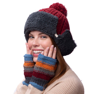 A person with long hair is wearing a red and gray winter hat with ear flaps, and a pair of Short Cuff Handwarmers that feature multicolored handmade patterns in shades of blue, orange, and red. The person's hands are touching their cheeks against a plain white background.